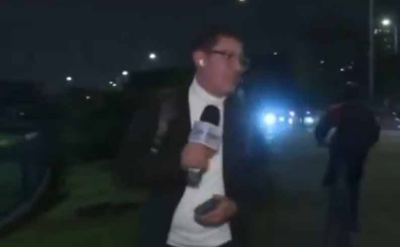 Mann mit Brille und Mikrofon steht nachts an einer Straße und weicht zur Seite aus, im Hintergrund läuft eine Person, ein Fahrzeug leuchtet blau.