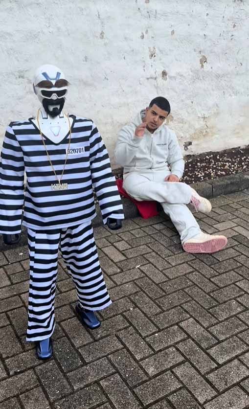 Eine Roboter in Sträflingskostüm mit Maske und Goldkette steht neben einem sitzenden AgaaMuza im hellen Jogginganzug vor einer Wand.