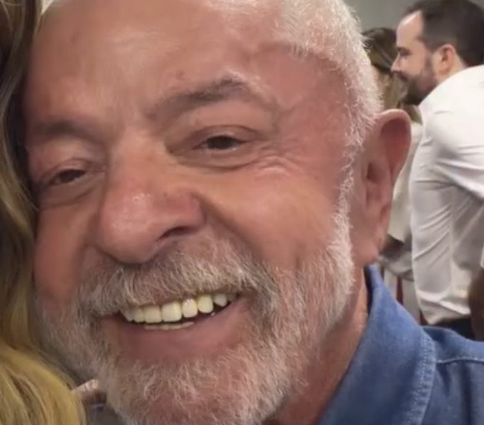 Homem idoso sorridente, de cabelo e barba brancos, com camisa jeans, ao lado de uma pessoa de cabelos loiros encostada em seu rosto