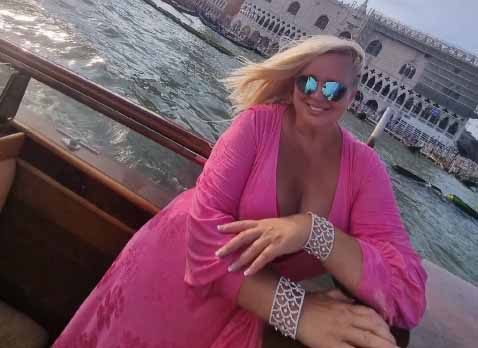 Mulher loira de óculos escuros espelhados e vestido rosa apoia-se sorrindo na amurada de um barco diante de um canal.