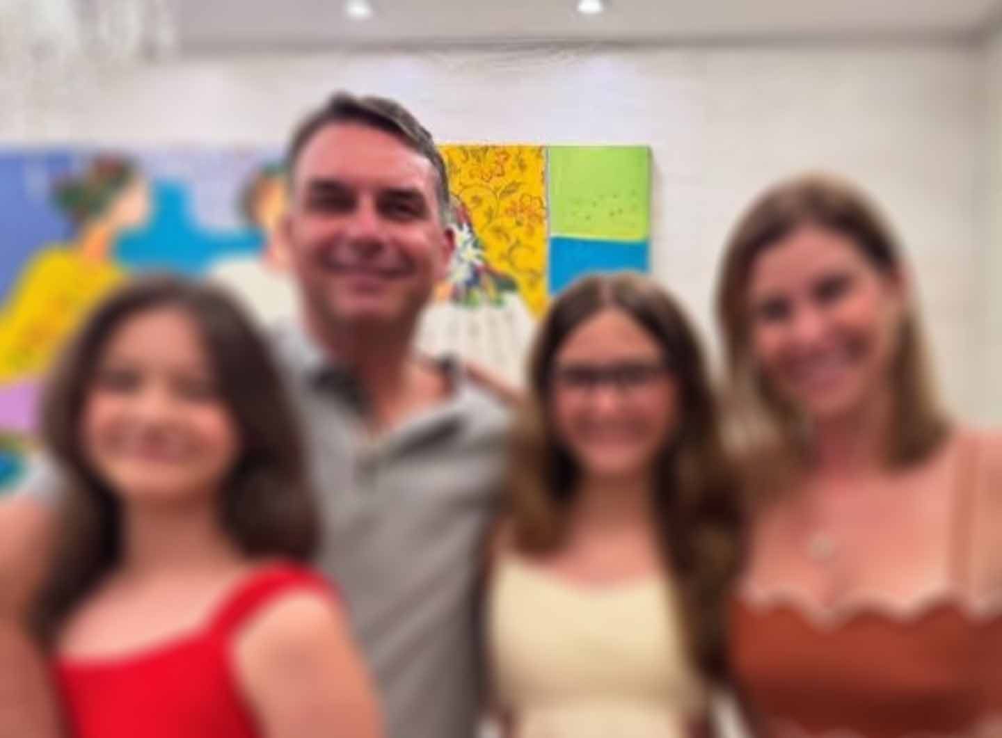 A man in a gray polo shirt stands smiling with two girls and a woman in front of colorful paintings in a room; the image is blurry.