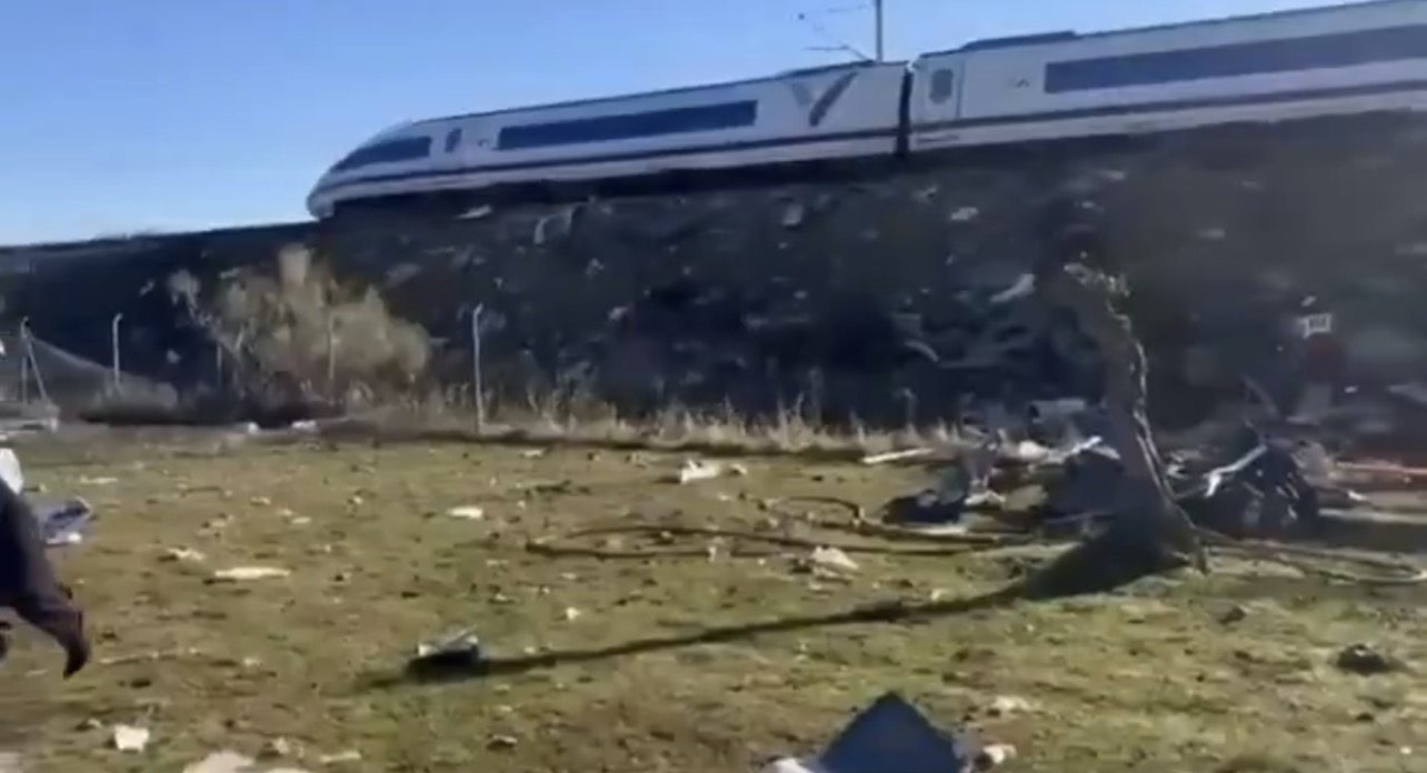 White high-speed train runs along an embankment; debris and wreckage lie scattered across a meadow in the foreground.