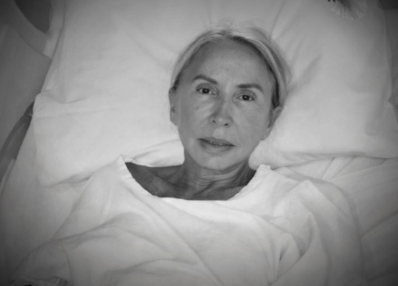 Black-and-white photo of a woman with light, combed-back hair lying on a pillow in a bed and looking into the camera