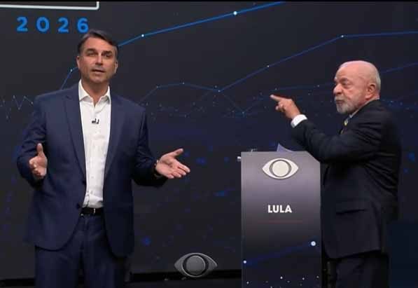 Zwei Männer in dunklen Anzügen stehen auf einer Fernsehbühne; einer breitet die Arme aus, der andere zeigt neben einem Pult mit Aufschrift „LULA“ auf ihn.