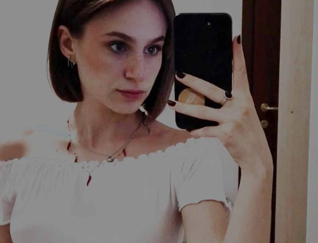 Jeune femme aux cheveux bruns coupés en carré au menton, vêtue d'un chemisier blanc à épaules dénudées, se photographiant dans un miroir avec un smartphone.