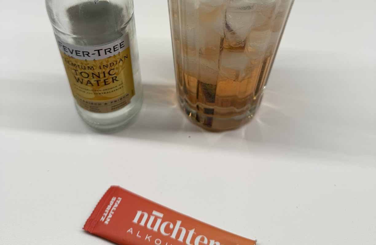 フィーバーツリーのトニックウォーターのボトルと、氷と琥珀色の飲み物が入ったグラス。手前にオレンジ色の小袋「nüchtern」