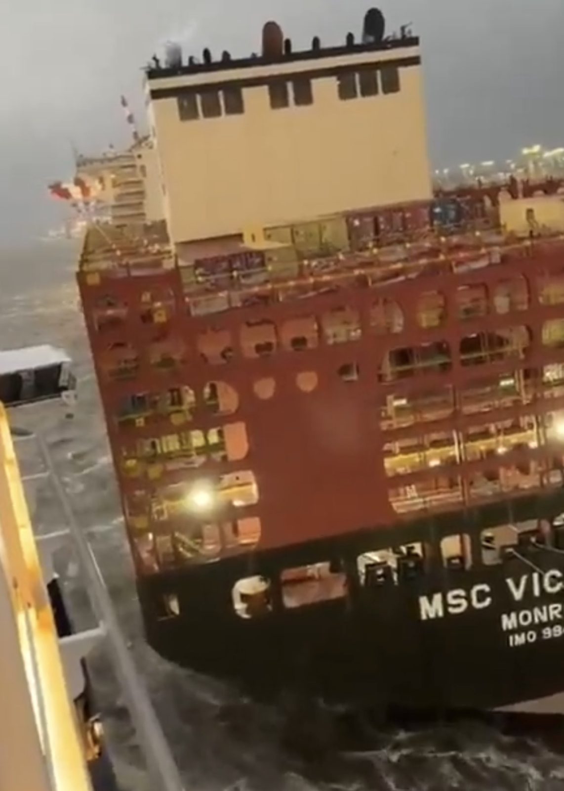 Navio porta-contêineres de casco vermelho sob céu escuro e mar agitado, com a inscrição "MSC Vic…" na popa e luzes acesas